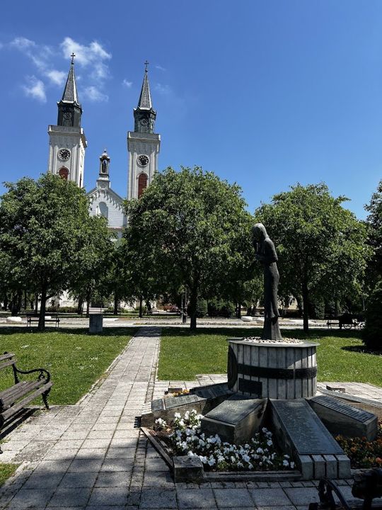 A memorial statue in Park heroja in Sombor, the twin towers of the Carmelite church behind the trees