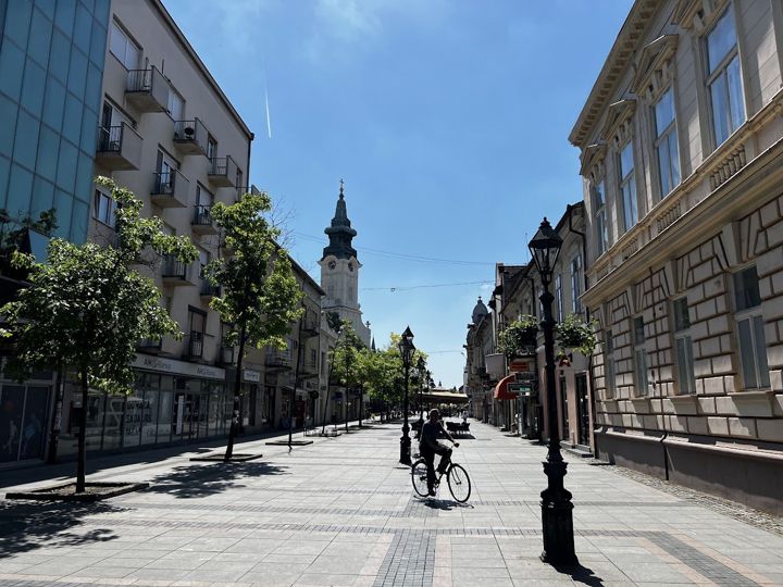 The pedestrian zone of Sombor: old façades, street lamps, a cyclist and a church tower at the end of the street