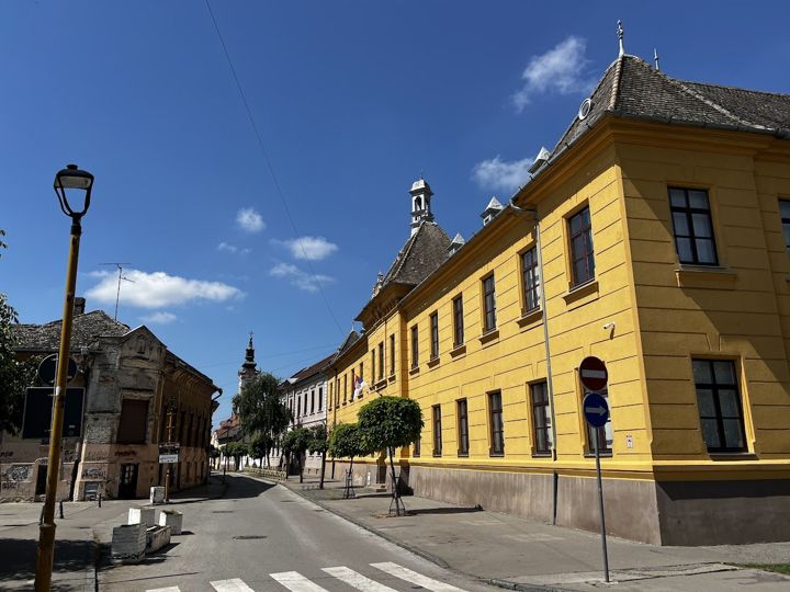 A long yellow building with roof turrets on a quiet street in Sombor, a church tower in the distance